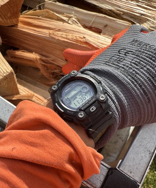 Schwarze Casio G-Shock mit Digitalanzeige am Handgelenk, im Hintergrund gespaltenes Brennholz und ein grauer Arbeitshandschuh